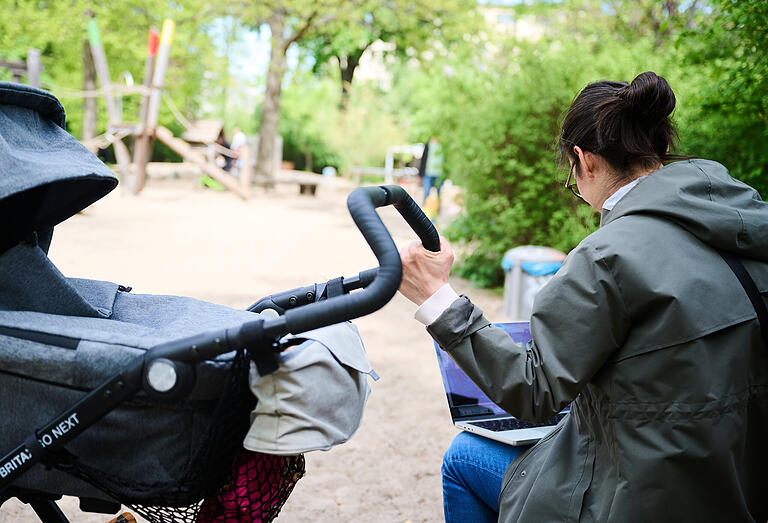 Alles gleichzeitig im Griff? Das Bild zeigt eine Frau mit Kinderwagen und Laptop auf dem Spielplatz.