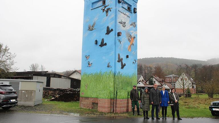 Landrätin Sabine Sitter, Bürgermeister Dirk Schiefer sowie Angehörige von Peter Paul vor dem frisch bemalten Herzensprojekt 'Peter-Paul-Turm' in Mittelsinn. Den Turm zieren nun Vögel, Wolken und Grasmotive.