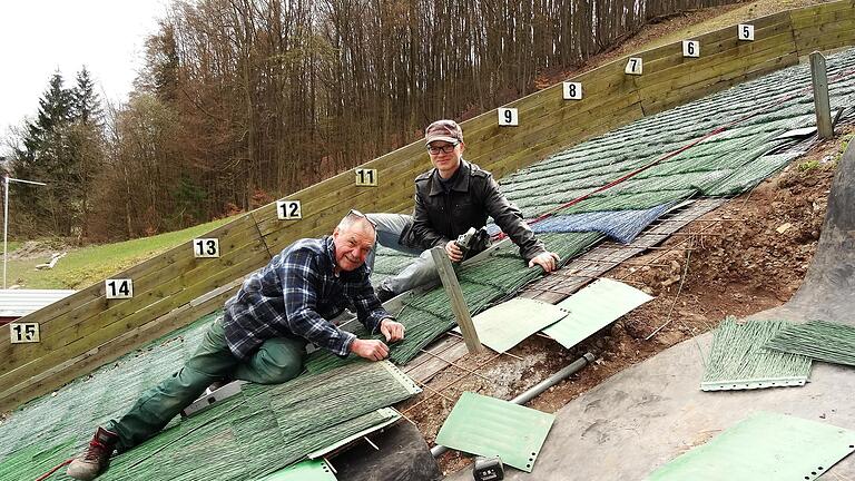 Beim Austauschen der Matten am Aufsprunghügel müssen sich Hans Beck (links) und sein Helfer Maximilian Lange am Steilhang festhalten.  Foto: Barbara Enders       -  Beim Austauschen der Matten am Aufsprunghügel müssen sich Hans Beck (links) und sein Helfer Maximilian Lange am Steilhang festhalten.  Foto: Barbara Enders