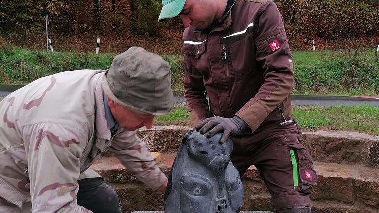 Die Gemeindearbeiter Berthold Schaab und Michael Detsch befestigen den von Gerwin Kellermann gemeißelten Rucksack aus Basaltstein.       -  Die Gemeindearbeiter Berthold Schaab und Michael Detsch befestigen den von Gerwin Kellermann gemeißelten Rucksack aus Basaltstein.