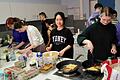 Schülerinnen und Schüler beim Zubereiten des French Toast und beim Schnippeln des Obstes, im Hintergrund werden Pancakes und Baked Beans zubereitet sowie Würstchen gebraten.