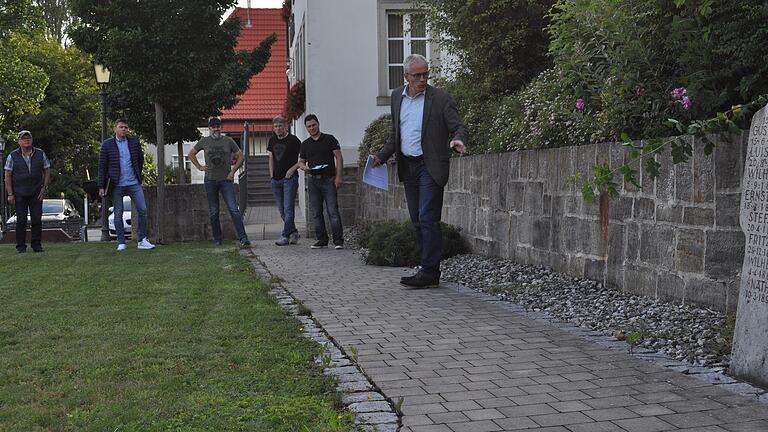 Ermershausens Bürgermeister Günter Pfeiffer (rechts) zeigt den Gemeinderäten zwischen dem Rathaus und dem Ehrenmal den Standort für den Koffer im Rahmen des Projekts Denkort Aumühle.