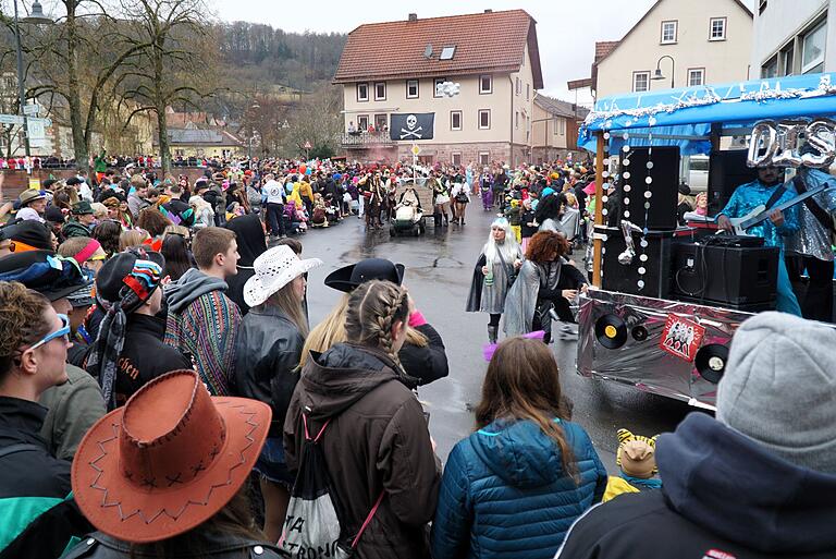 Der Faschingszug am Dorfplatz zwischen Kriegerdenkmal und Schondrabrücke, gesäumt von Zuschauern.