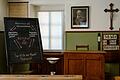 Das Klassenzimmer um 1910 weist auf den hierarchischen Aufbau hin: die Bänke auf unterster Ebene sind nach vorne ausgerichtet, erhöht auf einem Podest das Lehrerpult. Das zeigt den Ausdruck der Autorität des Lehrers als Vertreter der Staatsgewalt ...       -  Das Klassenzimmer um 1910 weist auf den hierarchischen Aufbau hin: die Bänke auf unterster Ebene sind nach vorne ausgerichtet, erhöht auf einem Podest das Lehrerpult. Das zeigt den Ausdruck der Autorität des Lehrers als Vertreter der Staatsgewalt mit Strafbefugnis; über dem Lehrer deutlich sichtbar für die Schüler hängen das Bild des Landesmonarchen (Prinzregent Luitpold) und das Kruzifix: Die Schüler sollen stets vor Augen haben, wem sie Gehorsam schuldig sind, das Bild des Monarchen soll sich einprägen.