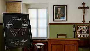 Das Klassenzimmer um 1910 weist auf den hierarchischen Aufbau hin: die Bänke auf unterster Ebene sind nach vorne ausgerichtet, erhöht auf einem Podest das Lehrerpult. Das zeigt den Ausdruck der Autorität des Lehrers als Vertreter der Staatsgewalt ...       -  Das Klassenzimmer um 1910 weist auf den hierarchischen Aufbau hin: die Bänke auf unterster Ebene sind nach vorne ausgerichtet, erhöht auf einem Podest das Lehrerpult. Das zeigt den Ausdruck der Autorität des Lehrers als Vertreter der Staatsgewalt mit Strafbefugnis; über dem Lehrer deutlich sichtbar für die Schüler hängen das Bild des Landesmonarchen (Prinzregent Luitpold) und das Kruzifix: Die Schüler sollen stets vor Augen haben, wem sie Gehorsam schuldig sind, das Bild des Monarchen soll sich einprägen.