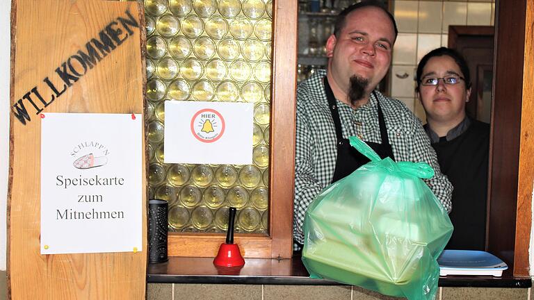 Mirko und Katharina Hauptig vom Gasthaus 'Schlappn' in Gerolzhofen reichen das typische Essen-zum-Mitnehmen-Paket durch das Fenster am Eingang ihrer Gaststätte. Mirko und Katharina Hauptig vom Gasthaus 'Schlappn' in Gerolzhofen reichen das typische Essen-zum-Mitnehmen-Paket durch das Fenster am Eingang ihrer Gaststätte.