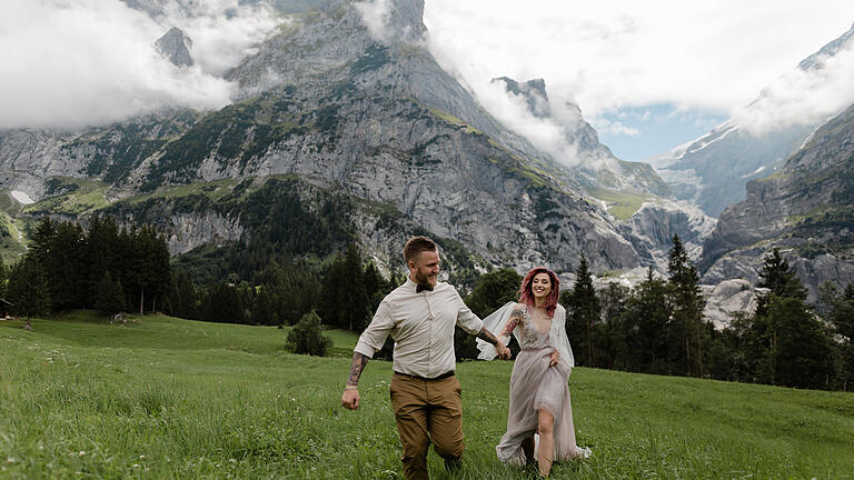 happy bride and groom holding hands and walking on alpine meadow with clouds       -  Heiraten auf einer Alm - ein Mädchentraum wird wahr! Trotzdem gibt es auf tausenden Metern einiges zu beachten, damit die Berghochzeit nicht zum Albtraum wird.