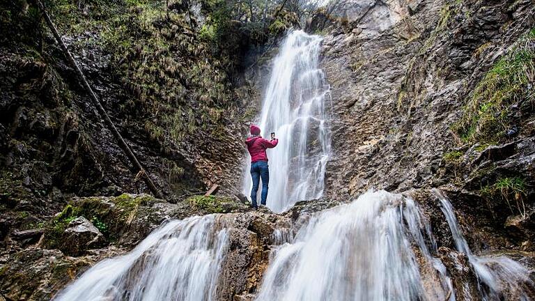 Eine Frau steht bei regnerischem Wetter am Wasserfall und nimmt ein Foto mit ihrem Smartphone auf.jpeg       -  Für Erlebnisse in freier Wildbahn gibt es spezielle Outdoor-Smartphones.