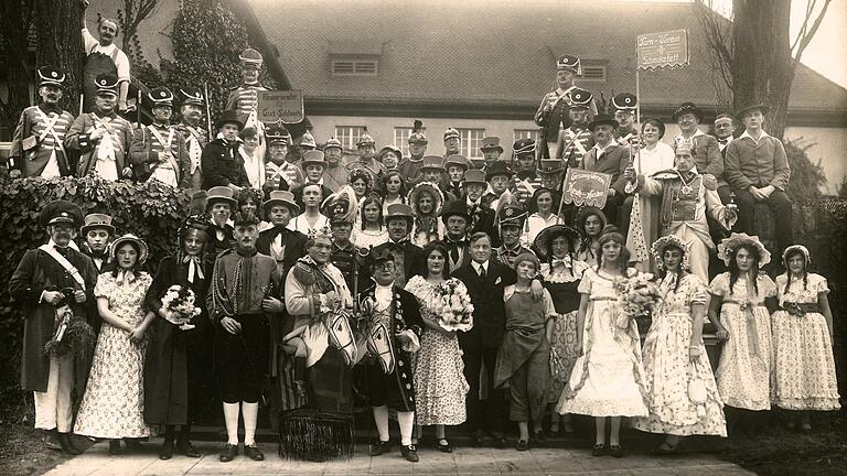 Zur Faschingszeit war es üblich, auf historische Themen und Ereignisse zurückzugreifen. Besonders beliebt waren so genannte „Lebende Bilder“, die unter einem solchen Motto standen. Auf diesem Foto aus dem Jahr 1928 des Lohrer Gesangsvereins 1843 mit Teilnahme der Freiwilligen Feuerwehr und des Turnvereins ist alles im Stil der Biedermeierzeit gehalten. Am linken oberen Bildrand sind Lohrer Bürger als Landwehr-Soldaten in der napoleonischen Zeit kostümiert und geben einen ungefähren  Eindruck, wie man sich einen Landwehrmann der damaligen Zeit vorstellen muss. Besonders die topfförmigen Kopfbedeckungen, die Tschakos, erinnern an die Lohrer Landwehr. Das Bild wurde vor dem Festsaal der Heil- und Pflegeanstalt aufgenommen. Er war noch bis in die Nachkriegszeit der einzige größere Saal in Lohr am Main. Dort wurde die „Faschingsoperette“ wohl auch aufgeführt.