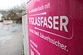Der Glasfaserausbau durch die Telekom in Lohr ist nach der Winterpause wieder angelaufen. Im Bild ein Verteilerkasten in Wombach mitsamt Werbeplakat der Telekom, im Hintergrund eine Glasfaserbaustelle.