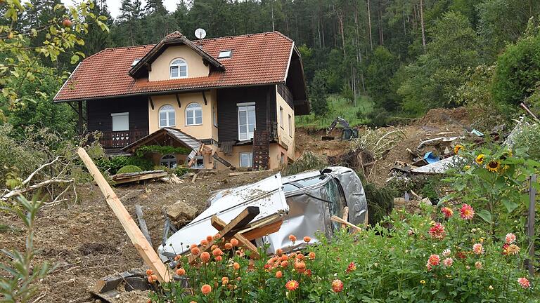 Nach Unwettern in Österreich.jpeg       -  Nach einem neuen Erdrutsch am Wochenende in Kärnten liegt dieses Auto unweit eines Hauses auf dem Dach, während im Hintergrund Aufräumarbeiten mit einem Bagger stattfinden.