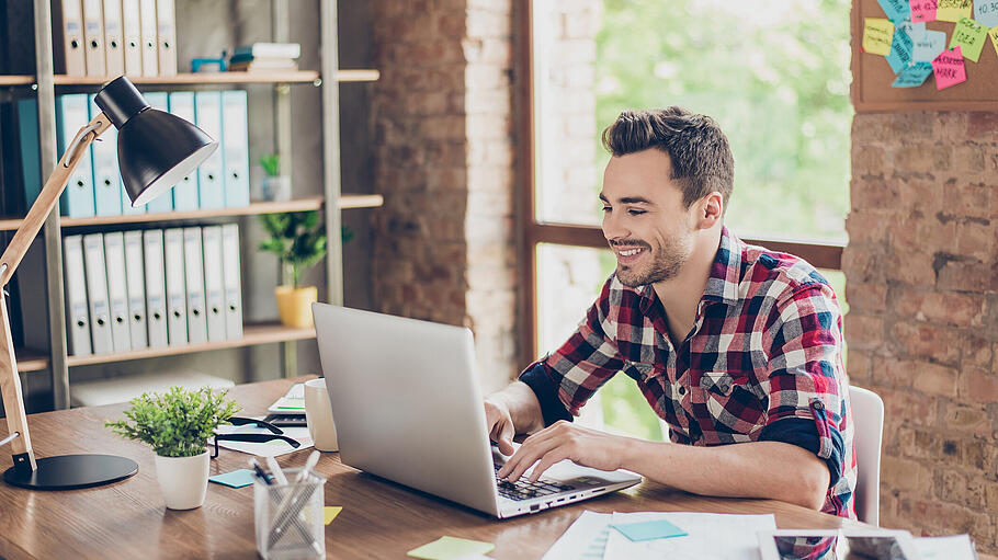 Cheerful young brunet freelancer is smiling, typing on his laptop in nice modern work station at home, in casual smart wear - Setzen Sie im Beruf Prioritäten, um Stress und Überstunden zu vermeiden. Cheerful young brunet freelancer is smiling, typing on his laptop in nice modern work station at home, in casual smart wear - Setzen Sie im Beruf Prioritäten, um Stress und Überstunden zu vermeiden.