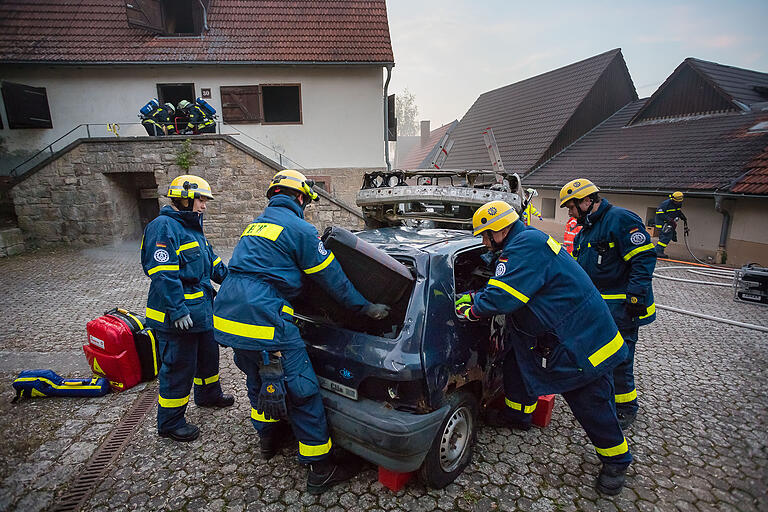 Während die Übungsteilnehmer des THWs sich um den Verkehrsunfall im Vordergrund kümmern und trainieren, dringen die Feuerwehrmänner im Hintergrund mit Atemschutzgerät in das simuliert brennende Haus ein. Während die Übungsteilnehmer des THWs sich um den Verkehrsunfall im Vordergrund kümmern und trainieren, dringen die Feuerwehrmänner im Hintergrund mit Atemschutzgerät in das simuliert brennende Haus ein.
