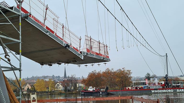 Am Samstagmorgen wurden auf der Margetshöchheimer Seite die ersten Fertigteile montiert. Wie auf dem Foto zu erkennen ist, werden die Anhängerseile zur Mitte der Tragseile hin immer kürzer. Damit wird sichergestellt, dass der Steg nach oben eine Wölbung erhält und so zu den beiden Seilen hin abfällt. In der Flussmitte fehlen die Anhänger völlig.  Hier wird ein Fertigteil direkt an die Tragseile montiert.