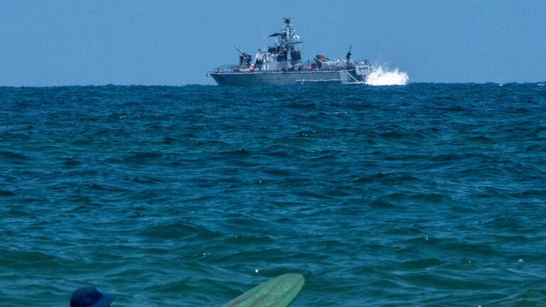 Israel       -  Ein Surfer wartet auf eine Welle, während ein israelisches Marineschiff im Mittelmeer vor der Küste von Hadera patrouilliert.