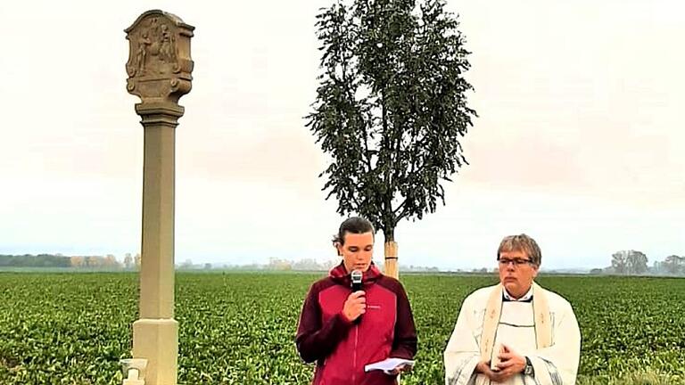 Pfarrer Amrehn segnet den wieder aufgerichteten Bildstock der Dreifaltigkeit aus dem Jahr 1787, Ilona Dusel erläutert die Bedeutung der Bildstöcke für das kulturell-künstlerische Leben in den Gemeinden und lädt die Kommunionkinder ein, die Inschrift zu entziffern - keine leichte Aufgabe für die Kinder.