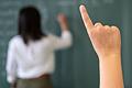 Lehrerin und Schüler.jpeg       -  Ein Schüler meldet sich, während die Lehrerin an die Tafel schreibt.