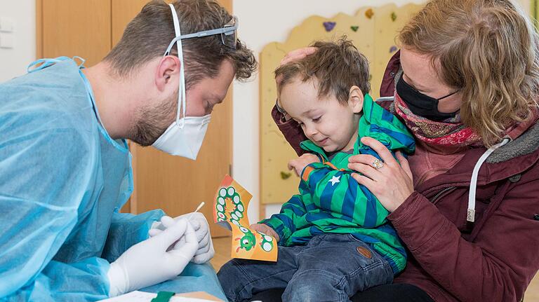 Assistenzarzt Philipp Fecher mit dem dreijährigen Tom, der auf dem Schoß seiner Mutter sitzt. Nach dem Corona-Test gibt es einen Belohnungsstempel ins Raupenheft.