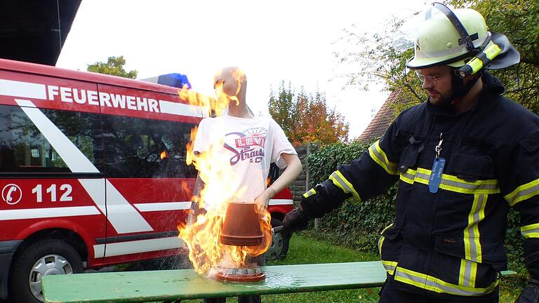 Moritz Hornung von der Volkacher Feuerwehr stellt den Brandausbruch in einem sogenannten Teelichtofen nach. Die Kleidung der im Hintergrund stehenden Puppe hat ebenfalls schon Feuer gefangen. Im Ernstfall könnte eine solche Situation in einem Wohnzimmer zur Katastrophe führen.