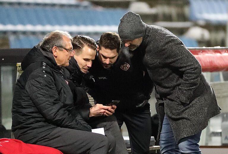 Teammanager Norbert Mahler, Vorstand Sport Sebastian Schuppan, Co-Trainer Philipp Eckart und Trainer Bernhard Trares schauen auf dem Smartphone die umstrittene Elfmeter-Situation.