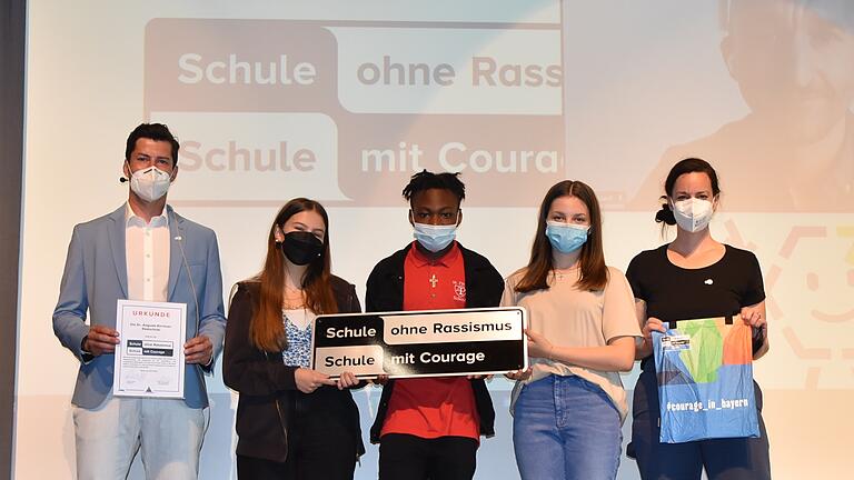 Die Dr.-Auguste-Kirchner Realschule (akr) Haßfurt wurde aufgenommen in das Netzwerk „Schule ohne Rassismus, Schule mit Courage“. Darüber freuen sich die beiden Projektleiter Roman Glöckner (links) und Vanessa Felber-Eschenfelder (rechts) sowie die Schülervertreter (ab zweite von links), Tessa Chatzilampros, Guy-Lennox Hely-Pelami und Nora Szmechel.  Im Hintergrund ist der Pate, der aus Zeil stammende Comedian Markus Barth, per Livestream zu sehen.