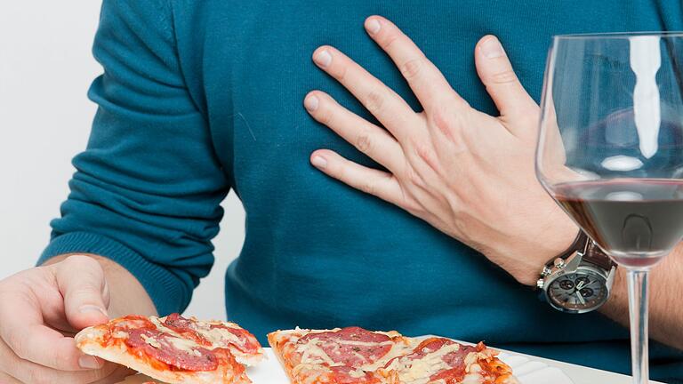 Mann isst Pizza und trinkt Rotwein - Fettiges Essen und Alkohol können Sodbrennen begünstigen. Mann isst Pizza und trinkt Rotwein - Fettiges Essen und Alkohol können Sodbrennen begünstigen.