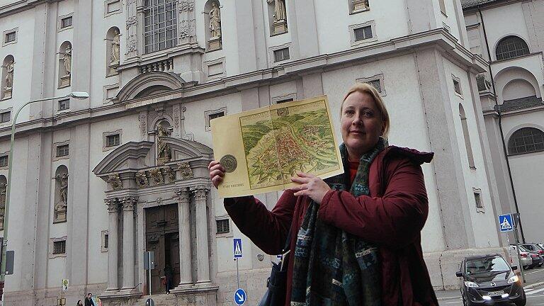 Claudia Jüngling steht vor Stift Haug und zeigt auf einer alten Karte von Würzburg, dass der Vorgängerbau bei der Stadtbefestigung im Weg war.