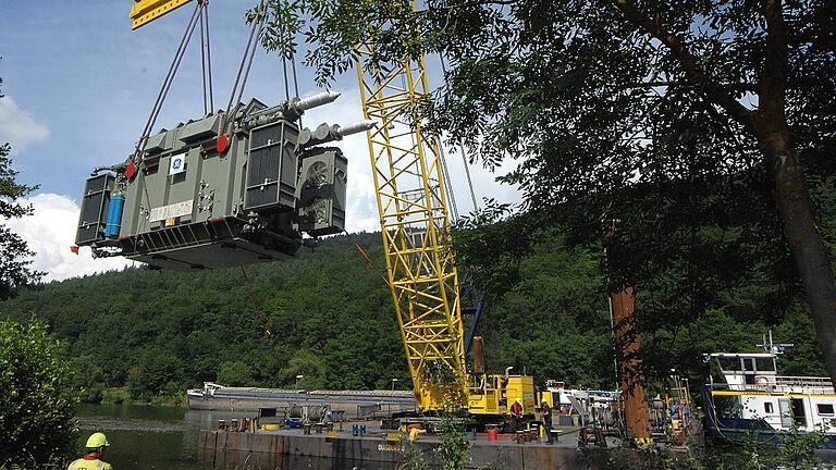 186-Tonnen-Transformator an den Stahlseilen des Schwimmkrans       -  (rp)   186 Tonnen &ndash; etwa das Gewicht von 135 VW Golf &ndash; hängen da an den Stahlseilen des Schwimmkrans: Am Mainufer bei Neuendorf wurde am Freitagnachmittag ein neuer Transformator für das Pumpspeicherkraftwerk Langenprozelten auf Spezialtransportfahrzeuge mit Rädern verladen. Wie Zahnräder greifen die Aufgaben der 80 beteiligten Spezialisten ineinander. Der Frachter, der den Trafo aus Mönchengladbach anlieferte, hat bereits wieder abgelegt, nachdem der Schwimmkran den Trafo hochgehievt hatte. Während die Fachleute der niederländischen Firma auf ihren nächsten Einsatz warten, schwitzen die Arbeiter der Transportfirma, die in sengender Hitze die stählernen Bodenplatten verlegen. Diese braucht es, damit die selbstfahrende, 23 Meter lange Transportplattform nicht im Boden einsinkt, wenn der Kran sich an Stahlseilen ans Ufer gezogen hat und seine Fracht ablässt. Auf einem Dutzend Achsen rollt diese Plattform dann aber nur 500 Meter weit bis zur Bundesstraße, wo der Trafo erneut umgeladen wird auf ein Spezialfahrgestell, das von zwei 650-PS-Maschinen bis zum Sindersbach gezogen und geschoben wird. Wegen der engen Kurven zum Kraftwerk hinauf kommt dann wieder die Transportplattform zum Einsatz. Das zweimalige Umladen war nötig, weil die Last wegen der Tragkraft der Brücke über die Eisenbahngleise bei Neuendorf auf mehr, nämlich 23 Achsen verteilt werden musste. Eine Million Euro kostet einer der Trafos, wobei der Transport mehr als zehn Prozent ausmacht. Im September wird der zweite Trafo ausgetauscht. Die beiden alten haben nach 40 Jahren ausgedient und werden verwertet.