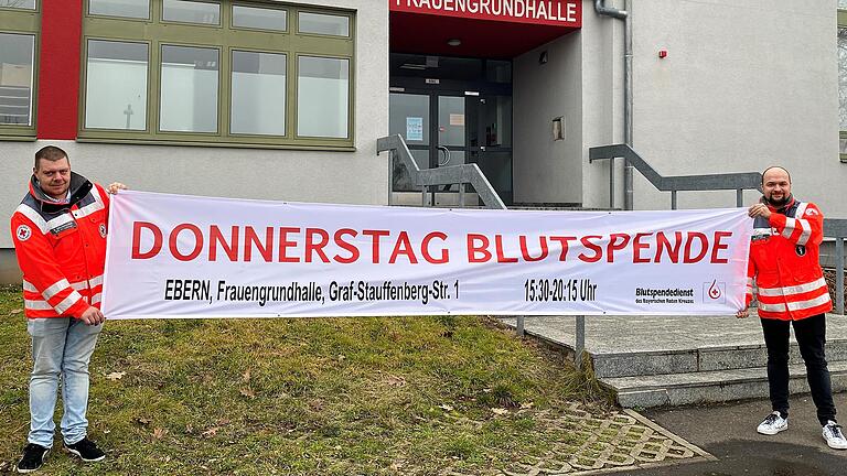 Um auf die neuen Termine für das Blutspenden in Ebern aufmerksam zu machen, wurden neue Transparente angeschafft, die in der jeweiligen Woche an verschiedenen Standorten in Ebern aufgehängt werden. Daniel Brunnhuber (links), stellvertretender Leiter der BRK-Bereitschaft Ebern, und Bereitschaftsmitglied Fabian Weber zeigen eines der Transparente vor dem Eingang zur Frauengrundhalle.