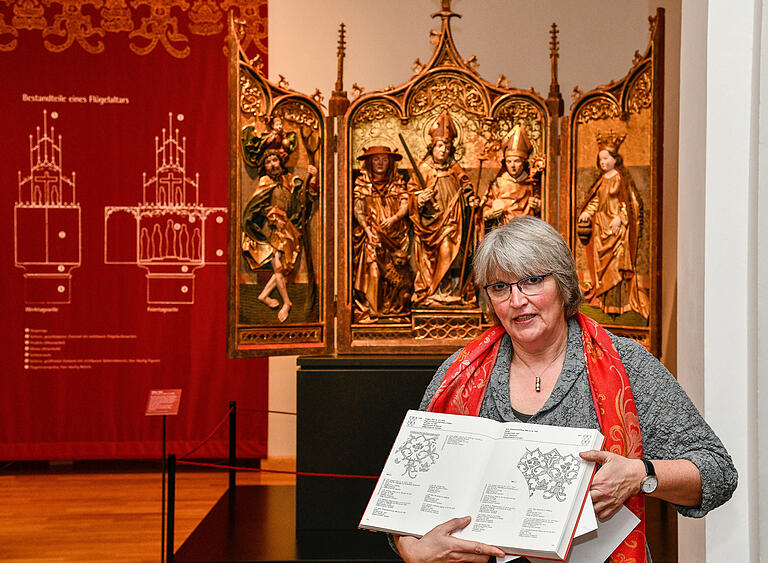 Claudia Lichte mit Musterbuch vor dem Kiliansaltar, der mit geöffneten Flügeln seine Festtagsseite zeigt.