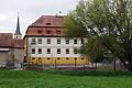 Die Rückseite des Rathauses in Prosselsheim mit einem Teil der Schlosswiese im Vordergrund und der Brücke mit ihrem Geländer über den Dettelbach (links). Hinter der 'Amtskellerei' soll einer der Eingänge zum neuen Naherholungsgebiet sein.