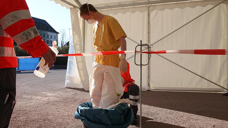 Nach dem Einsatz in der Teststelle brauchen die Mitarbeiter des Gesundheitsamts Hilfe, um die vielen Lagen Schutzkleidung abzulegen. Dazu stehen sie in einem flachen Abfalleimer, denn die Schutzkleidung muss als infektiöser Abfall aufwändig entsorgt werden.