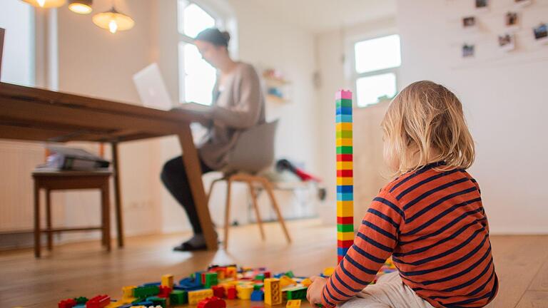 Ein Kind spielt im Vordergrund, arbeitende Person im Hintergrund.jpeg       -  Meist sind es Frauen, die zu Hause bleiben, um sich um die Kinder zu kümmern. Wie hoch fällt ihre Rente aus?