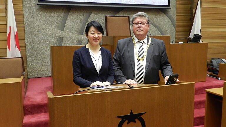 Bürgermeisterin Naomi Koshi und Bürgermeister Adolf Bauer am Rednerpult des Sitzungssaals mit Stadtwappen im Rathaus der Stadt Otsu.