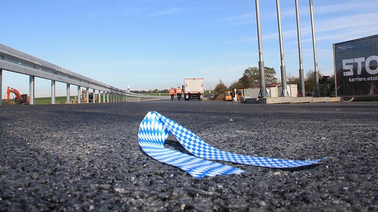 Nach der offiziellen Freigabe der Ortsumgehung von Mönchstockheim liegt ein Fetzen des symbolisch durchtrennten Stoffbandes auf dem Asphalt. Arbeiter machen sich im Hintergrund daran, letzte Hindernisse von der Fahrbahn zu räumen, bevor im Lauf des Freitagnachmittags die ersten Fahrzeuge auf die nagelneue Straße gelassen wurden.