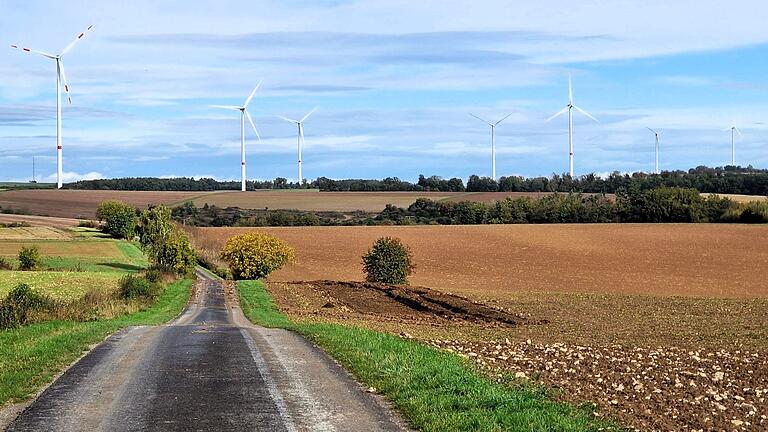 Auf dem Natura Trail führt der blaue Keil ab der Waldecke auf asphaltierter Straße über das offene Feld, im Hintergrund der Windpark Güntersleben. Auf dem Natura Trail führt der blaue Keil ab der Waldecke auf asphaltierter Straße über das offene Feld, im Hintergrund der Windpark Güntersleben.