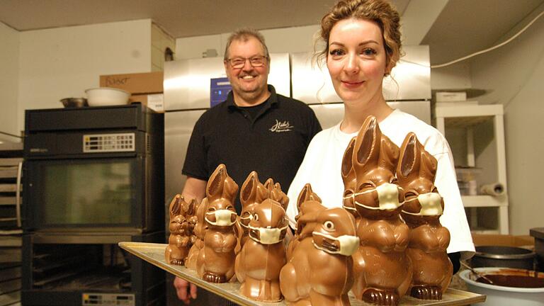 So sehen sie aus, die trotz Mundschutz lustig drein blickenden Schoko-Osterhasen aus der Backstube der Familie Häcker, die hier von Carola Häcker präsentiert werden. Im Hintergrund Konditormeister Herbert Häcker.&nbsp;