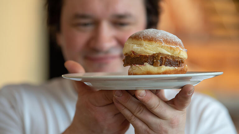 Neue Krapfen-Kreationen       -  Der Bäckermeister Florian Perkmann hält einen Krapfen, der mit Leberkäse und süßem Senf belegt wurde in der Hand.