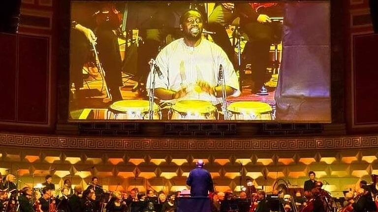 Hier trommelt Niang Serigne auf einer großen Bühne und unterstützt das Orchester mit seiner Kunst. Sein Foto wird auf einer großen Leinwand übertragen. Foto: Biggi Trost       -  Hier trommelt Niang Serigne auf einer großen Bühne und unterstützt das Orchester mit seiner Kunst. Sein Foto wird auf einer großen Leinwand übertragen. Foto: Biggi Trost