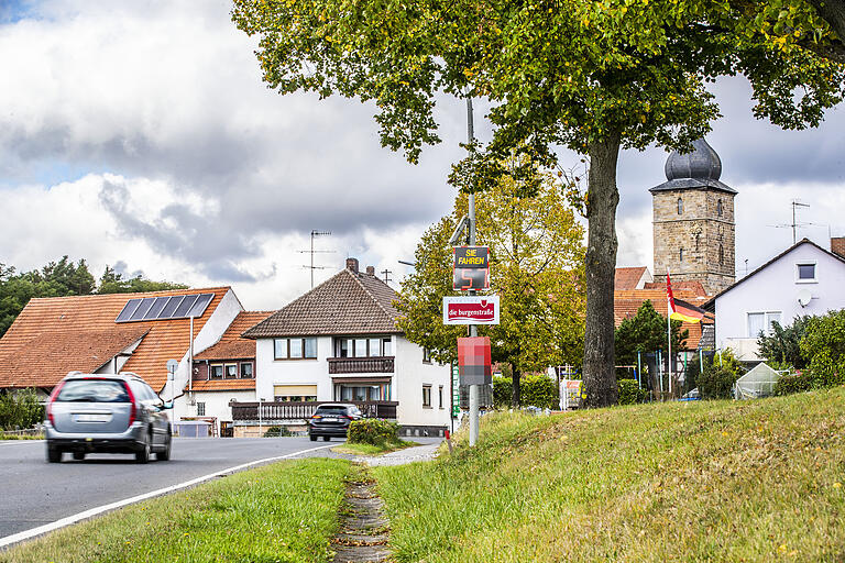 An der Ortsdurchfahrt von Pfarrweisach steht ein Geschwindigkeitsmesser und misst die Fahrgeschwindigkeit der Fahrzeuge auf der B279. Im Hintergrund sieht man die Dorfkirche.