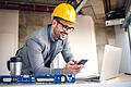Smiling attractive caucasian architect with helmet on head holding smart phone and looking at laptop while standing in building in construction process.       -  Die eVergabe-Plattform der Main-Post erleichtert den Vergabealltag im öffentlichen Sektor.