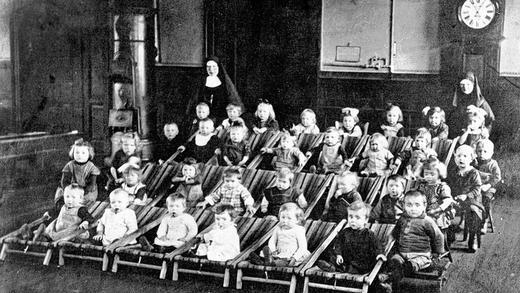 Zwischen Kanonenofen und Standuhr: Zwei Schwestern aus der Kongregation der Töchter des Allerheiligen Erlösers und Buben und Mädchen der damaligen Kinderbewahranstalt auf einem undatierten Foto. Die Kinder haben es sich auf Liegestühlen bequem gemacht. Zwischen Kanonenofen und Standuhr: Zwei Schwestern aus der Kongregation der Töchter des Allerheiligen Erlösers und Buben und Mädchen der damaligen Kinderbewahranstalt auf einem undatierten Foto. Die Kinder haben es sich auf Liegestühlen bequem gemacht.