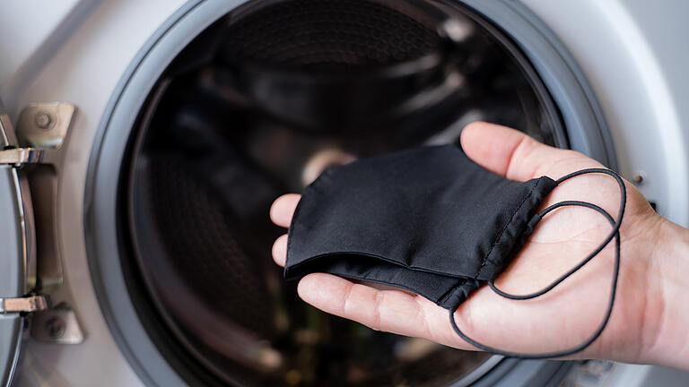 The hand of a man who is throwing a black mask into the washing machine       -  Nach dem Tragen sollen Masken regelmäßig gereinigt werden. Wie aber wird die Maske virenfrei?