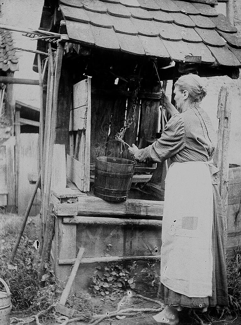 Frau beim Wasserholen am Brunnenhäuschen: Der Stadtarchivar verweist auf ihre Arbeitskleidung mit Schürze, die früher bei der Haus- und Gartenarbeit stets getragen wurde.