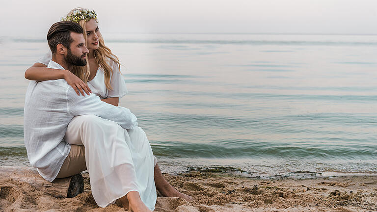 elegant groom and bride sitting on log on beach and looking away       -  Strandhochzeit, Leichtturm oder Hochzeitsmühle: Eine Hochzeit am Meer ist ein einzigartiges Erlebnis.