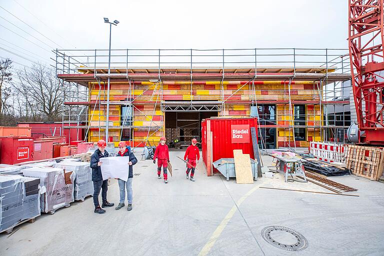 Vor der Baustelle stehen (von links) die beiden Bauleiter Julian Klier und Philipp Hoos, die beiden Azubis Vincent Hecht und Marcel Rüger tragen Eisenmaterial herbei.