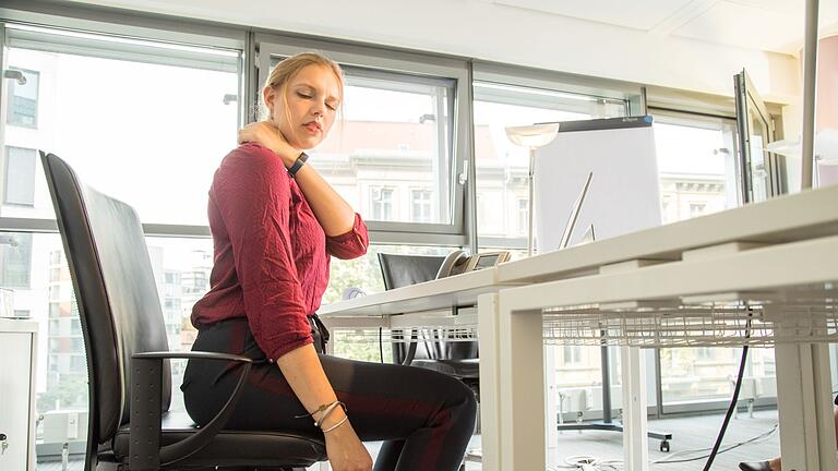 Eine Frau sitzt in einem Büro und hat Nackenschmerzen       -  Eine schiefe Körperhaltung, oft durch lange Büroarbeit verursacht, kann zu Verspannungen und Schmerzen führen.