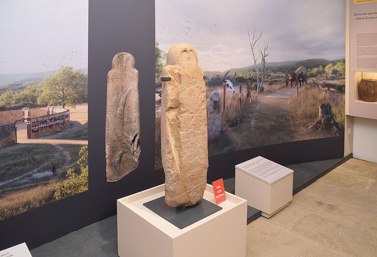 Der Menhir vor einem Panorama-Bild, auf dem die Landschaft aus der Zeit der Entstehung des Menhirs dargestellt ist. Links im Bild: der Bau der Kreisgrabenanlage bei Ippesheim.