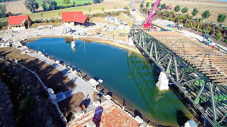 Am Donnerstag hievten zwei Schwerlastkräne die ehemalige Segnitzer Mainbrücke auf ihre endgütige Position über einem künstlich angelegten See bei Bolzhausen (Lkr. Würzburg). Die Brücke soll dort die Terrasse eines Wirtshauses werden, umgeben von einem Biergarten.