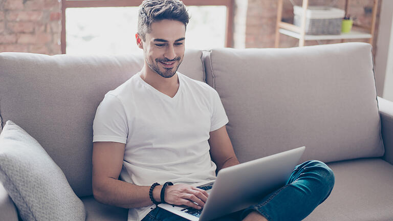 Young attractive smiling guy is browsing at his laptop, sitting at home on the cozy beige sofa at home, wearing casual outfit       -  Während der Corona-Krise gibt es viele digitale Wege, wie man regionale Händler und Restaurants unterstützen kann.